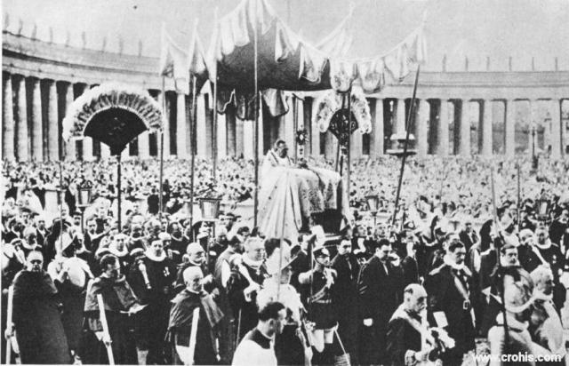 Tijelovska procesija u Rimu. Uz nova, velika i moderna okupljanja 30-tih godina 20. st. pozornost privlače i ona drevna, tradicijska, vjerska. O ovome svjedoči i mnoštvo na Trgu Sv. Petra tijekom tijelovske procesije. Slika je osobito važna jer prikazuje samog Svetog Oca, papu da predvodi obred. To se dogodilo prvi put poslije 1870. kada je talijanska vojska zauzela Rim, a papa se proglasio zatočenikom Vatikana. Odraz je poboljšanja odnosa između Svete stolice i talijanske države poslije sklapanja Vatikanskih ugovora 1929.